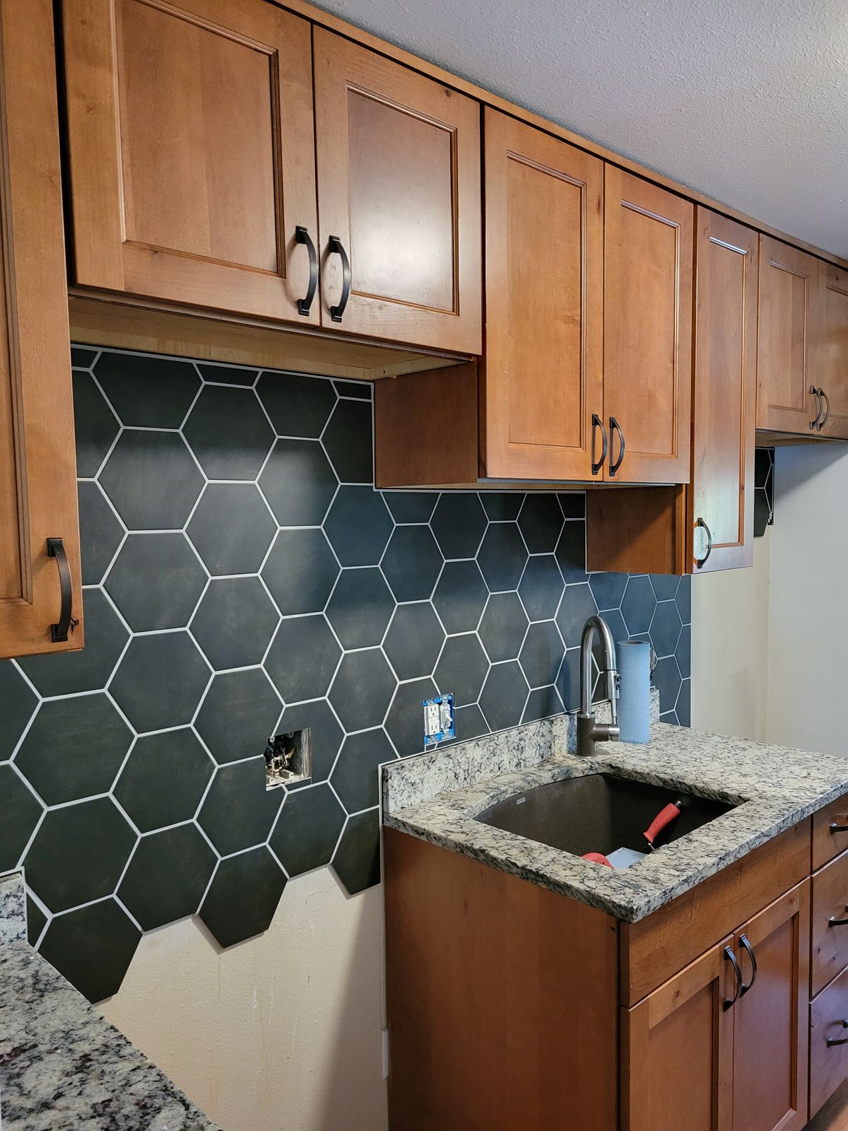 Kitchen with dark hexagonal tile backsplash installed but outlet cover plates missing, medium-wood cabinets, granite countertop with undermount sink, and a hand tool visible on the counter.