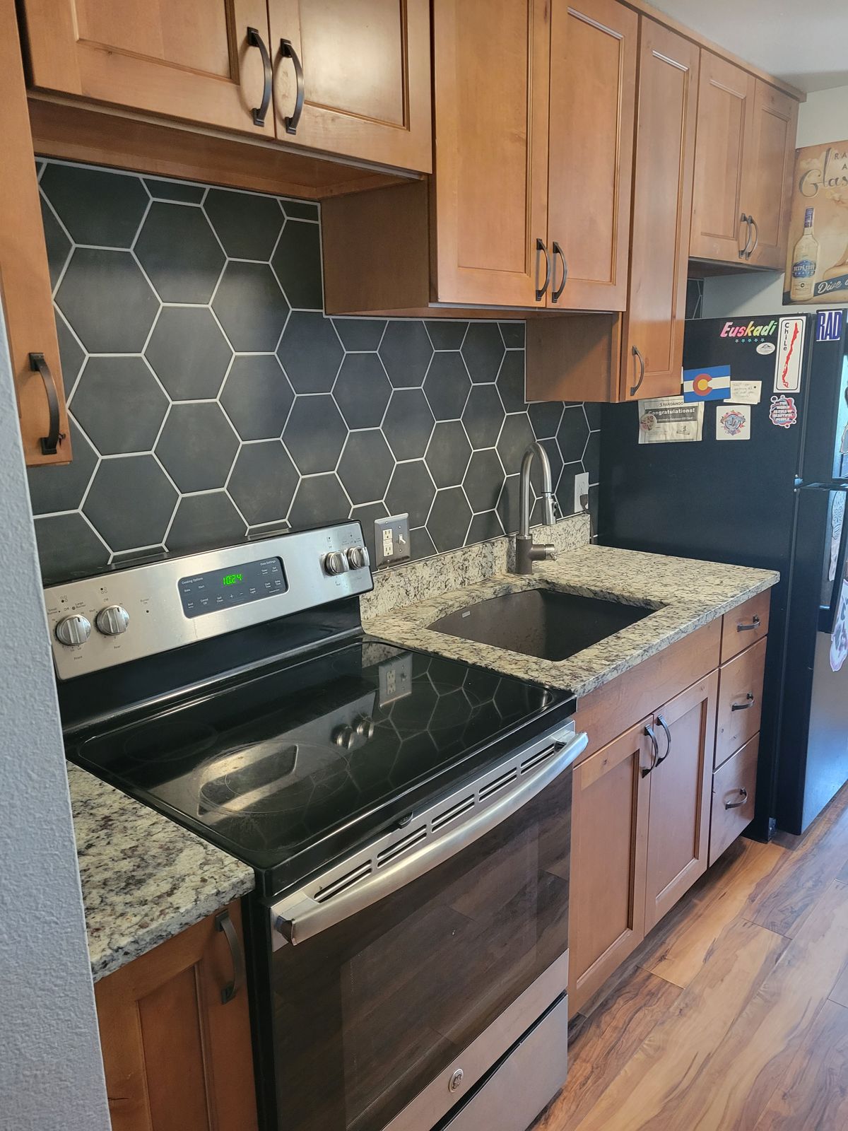 Finished kitchen showing dark hexagonal tile backsplash, medium-wood cabinets, granite countertop, undermount sink, stainless and black electric range, and wood-look flooring, with a refrigerator visible at the right edge.
