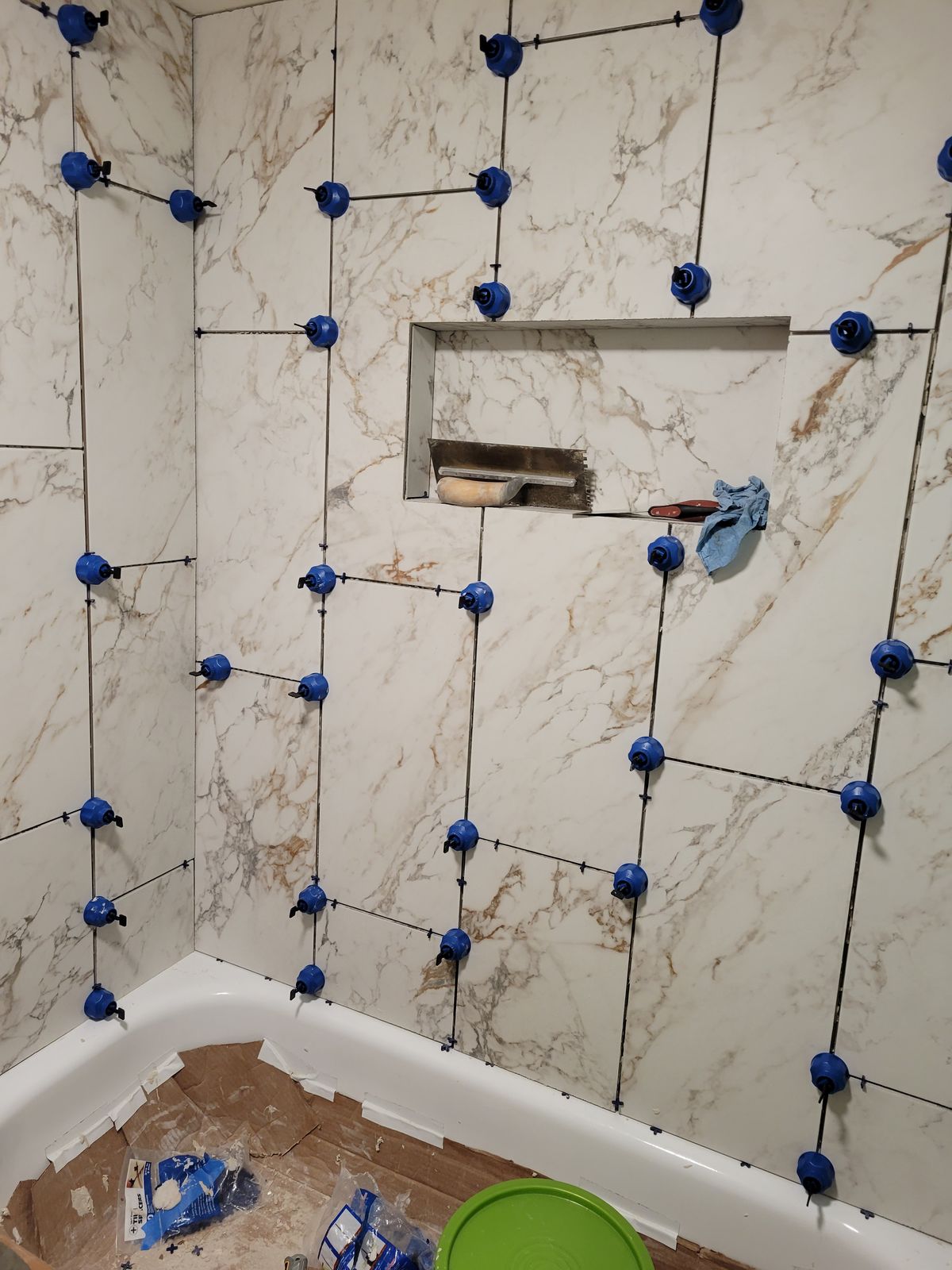 Tub surround with large-format marble-look tile set on three walls and blue leveling spacers still in place, recessed niche framed in metal, tub protected by cardboard, and tile tools visible on the floor.