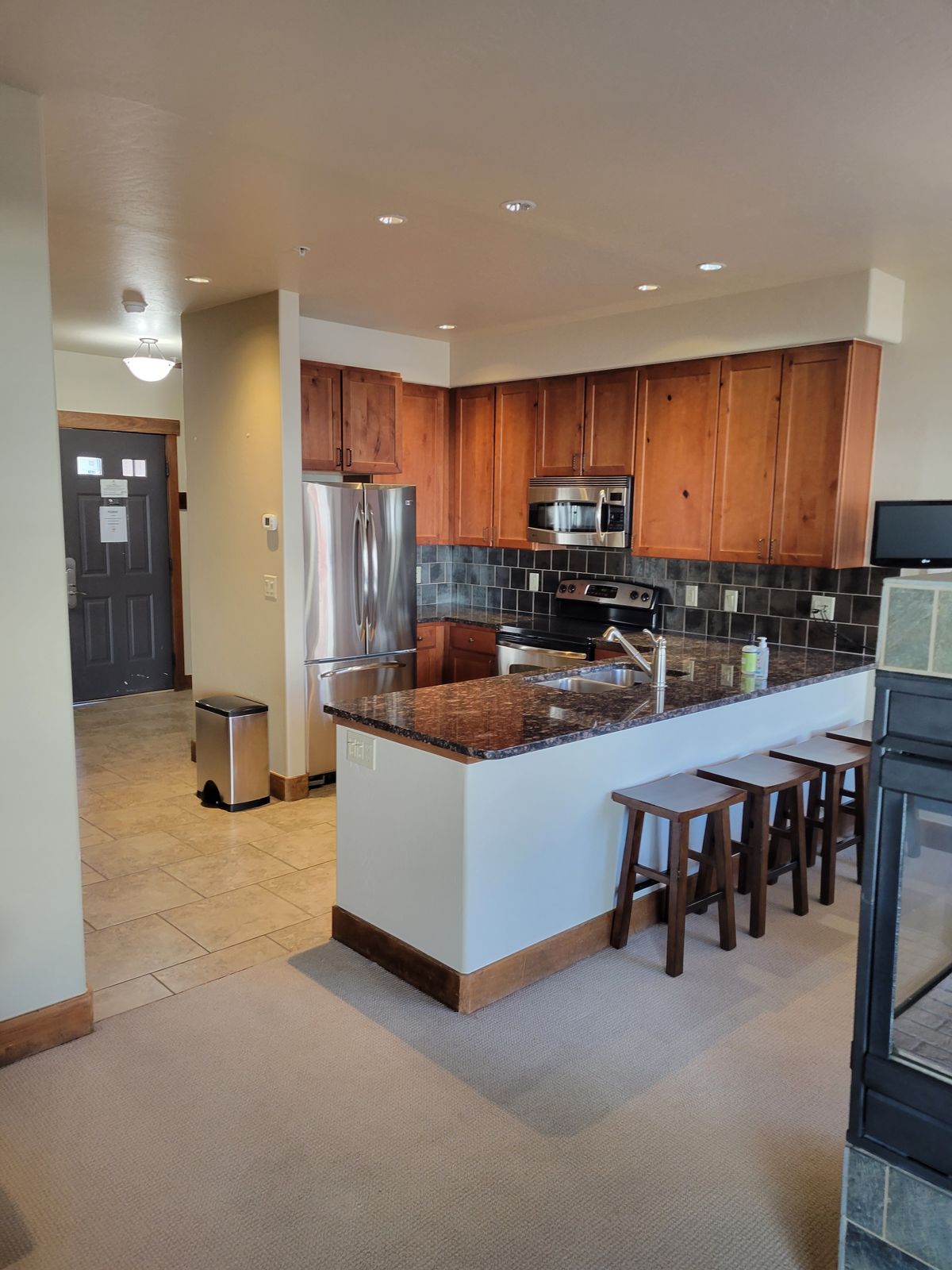 Finished kitchen with medium-toned wood cabinets, dark granite countertops, dark tile backsplash, stainless steel appliances, a peninsula with bar stools, and recessed lighting, with a front entry door visible in the background.