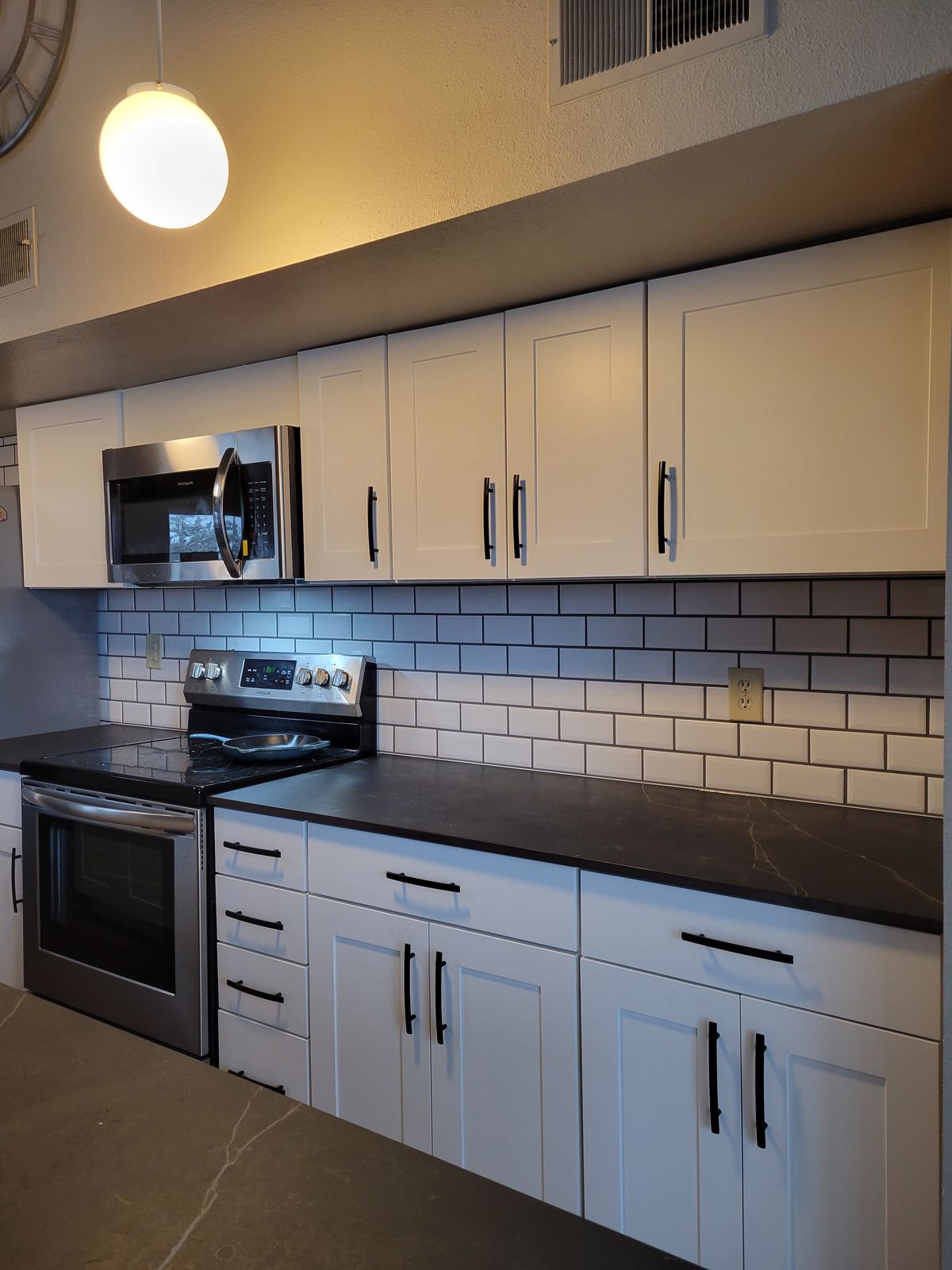 Completed kitchen run showing white shaker cabinets, white subway tile backsplash with dark grout, dark countertops, a stainless steel range and over-the-range microwave, and a globe pendant light overhead.