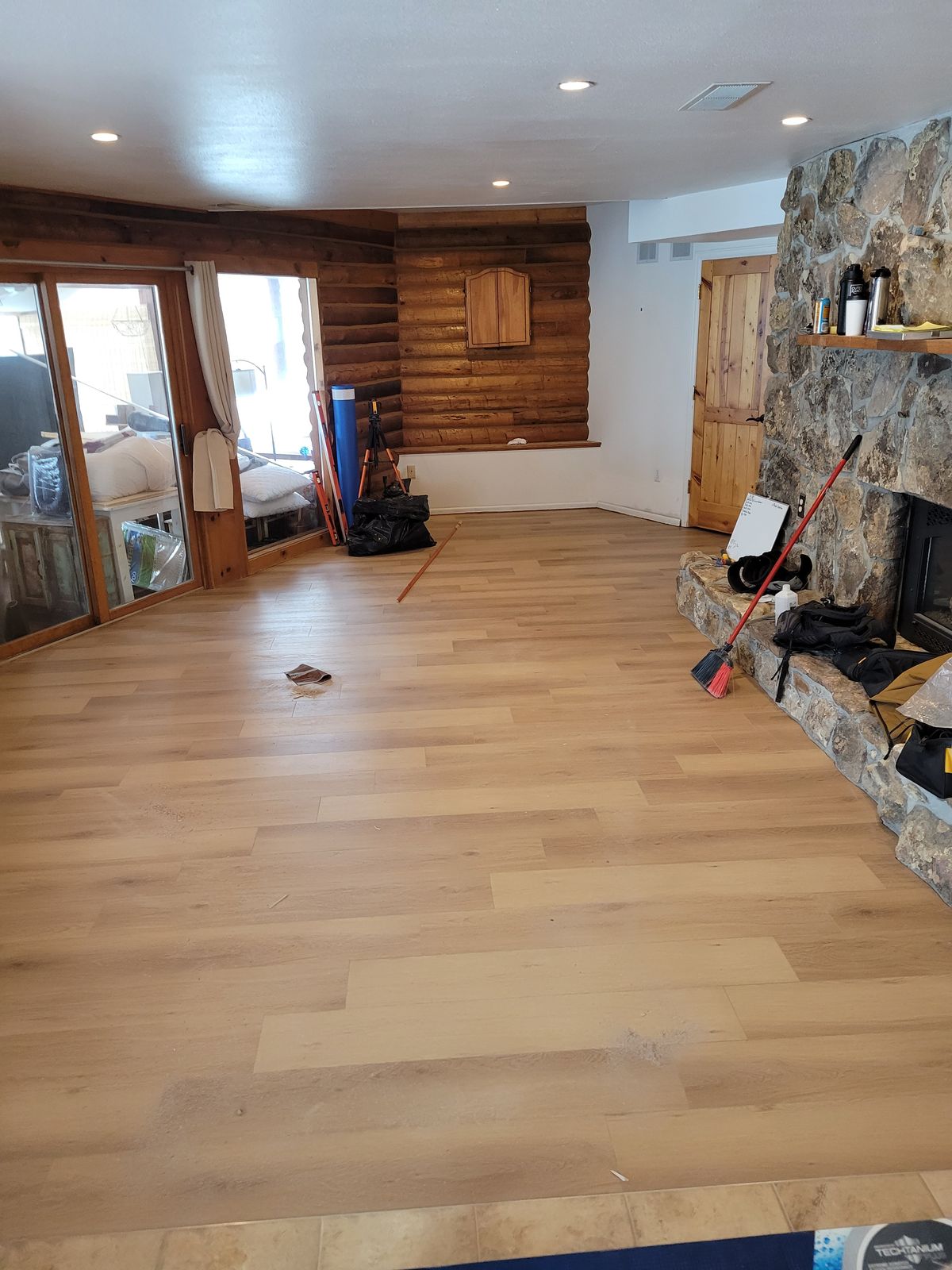Living room with new light-colored wood plank flooring laid across most of the floor, a stone fireplace on the right with a broom and work bags resting on the hearth, and a log-paneled wall with glass doors on the left.