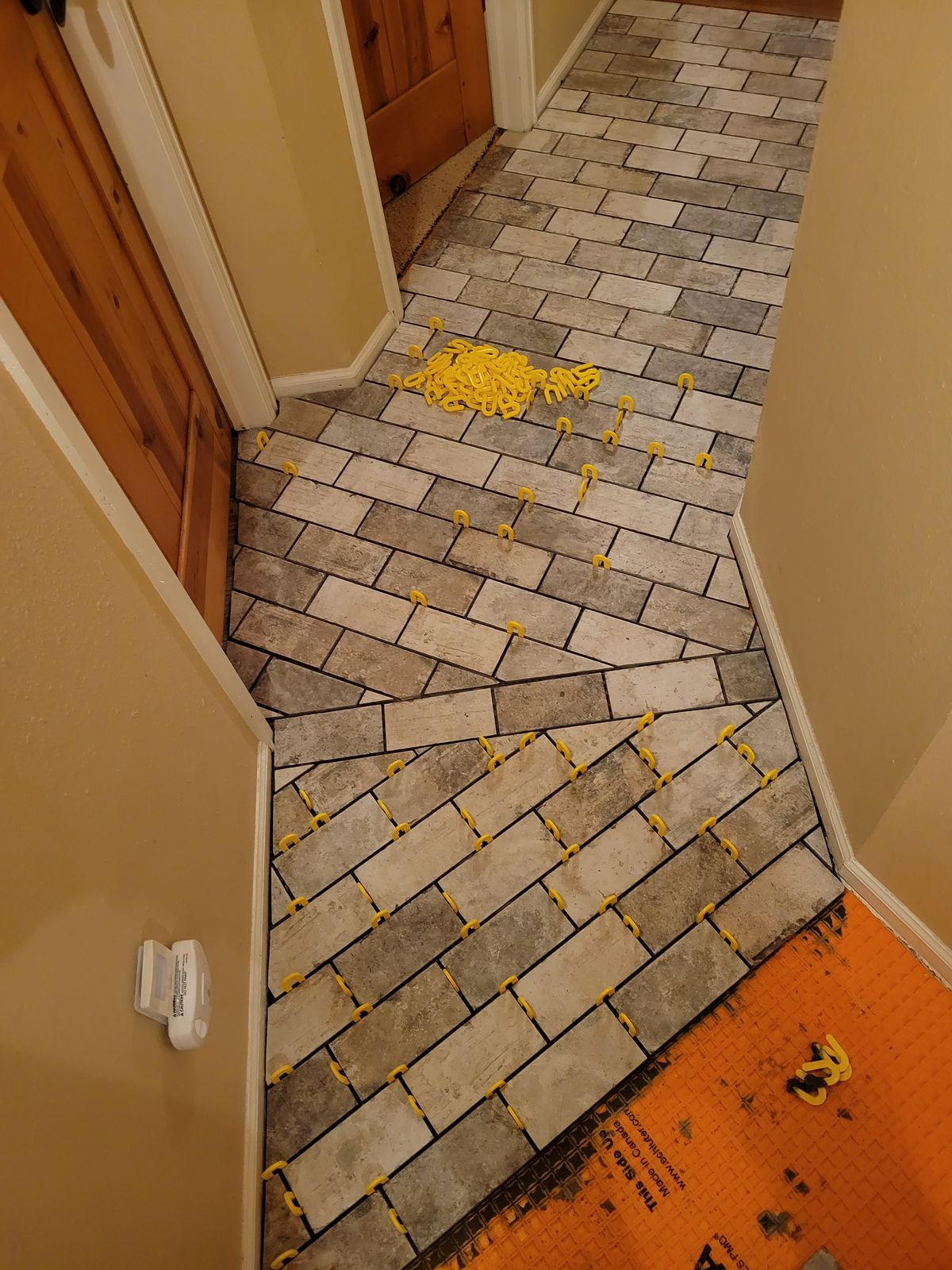 Hallway with diagonal-pattern tile flooring being set, yellow tile spacers still in place and orange underlayment visible at the near end.