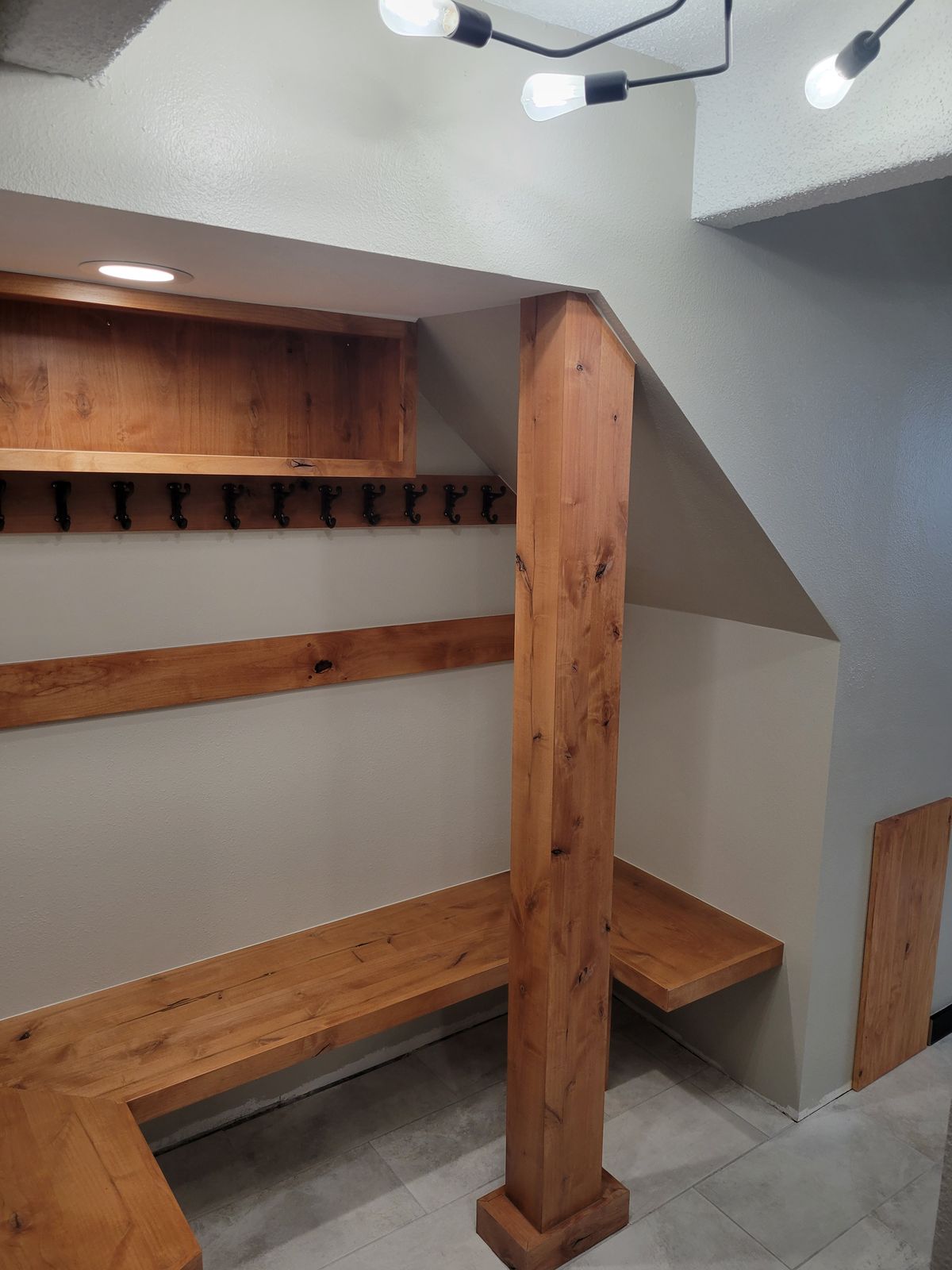 Finished under-stair mudroom nook with a knotty-wood L-shaped bench, upper shelf with coat hooks, a wood-wrapped post, tile flooring, and a multi-bulb pendant light.