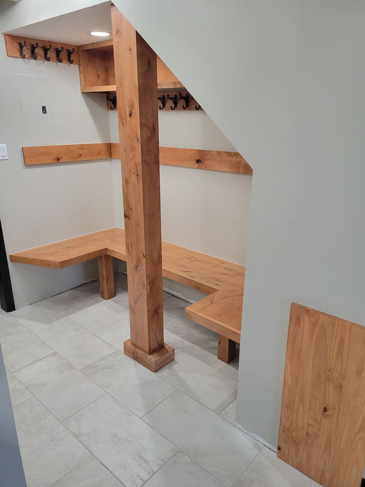 Finished mudroom under a staircase with natural wood built-in benches, shelves, and wall-mounted coat hooks over large-format tile flooring.