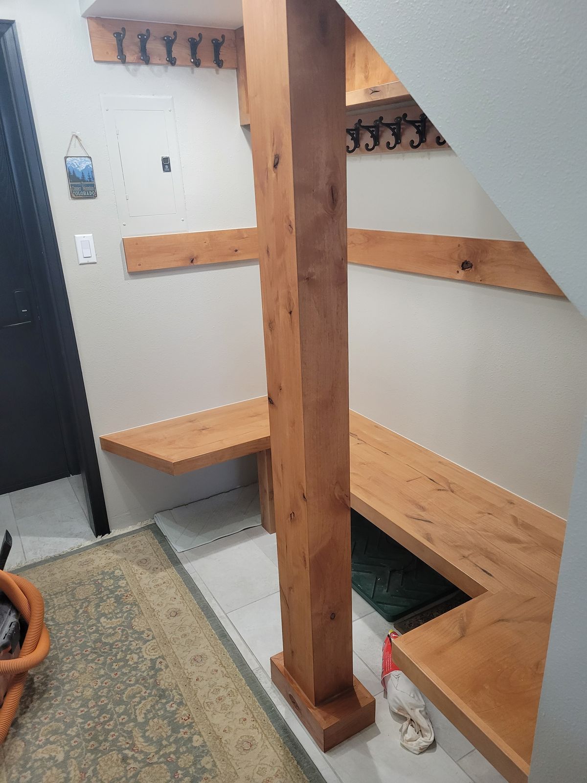 Wide view of the mudroom showing the central wood post, corner bench, shelving with coat hooks, a partial exterior door, and tile floor with an area rug; personal items visible on the bench.