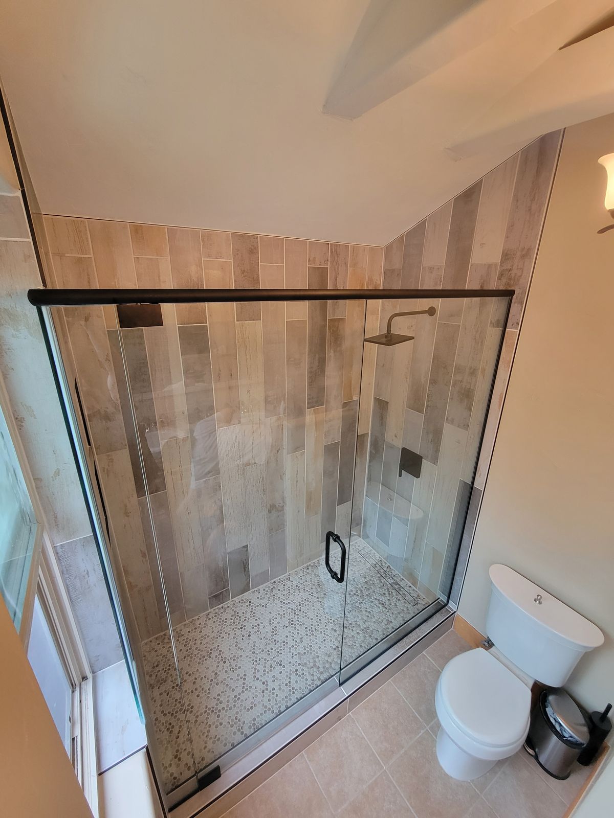 Finished bathroom featuring a frameless glass-panel shower enclosure with vertically stacked stone-look wall tile, a mosaic pebble floor, dark hardware, rain shower head, and a toilet visible beside it.