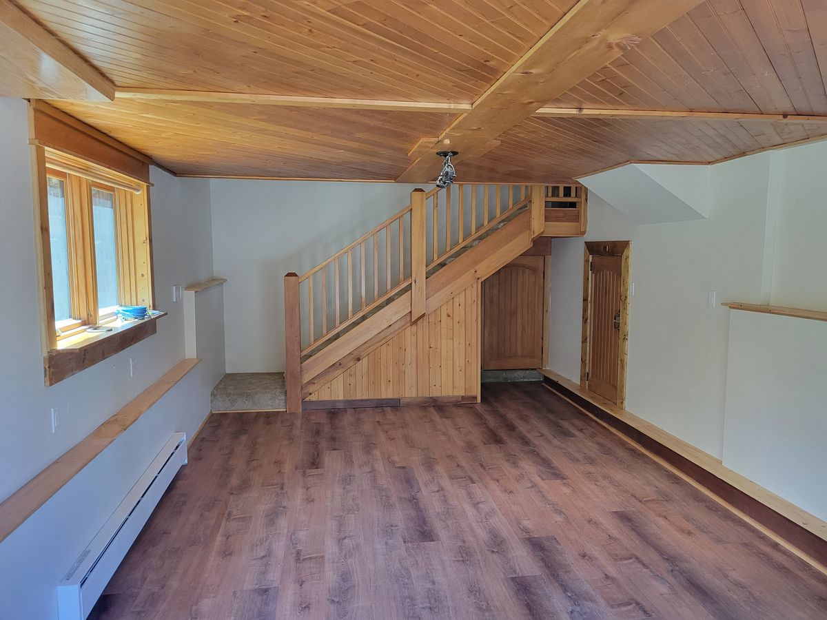 Empty living area with wood-plank laminate flooring, a wood-paneled staircase with wood railing, tongue-and-groove wood ceiling, and natural wood window and door trim.