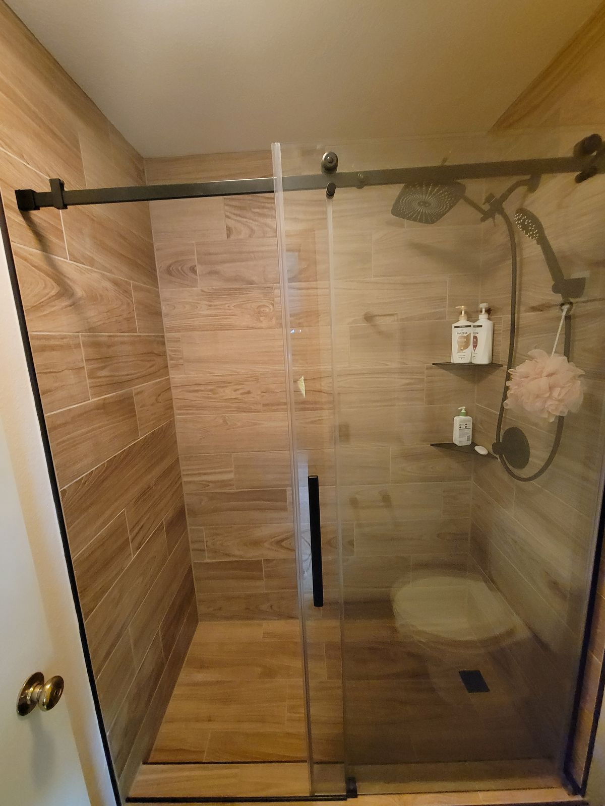 Finished walk-in shower with wood-look large-format tile on walls and floor, sliding frameless glass door with black hardware, built-in corner shelf, and handheld shower head.