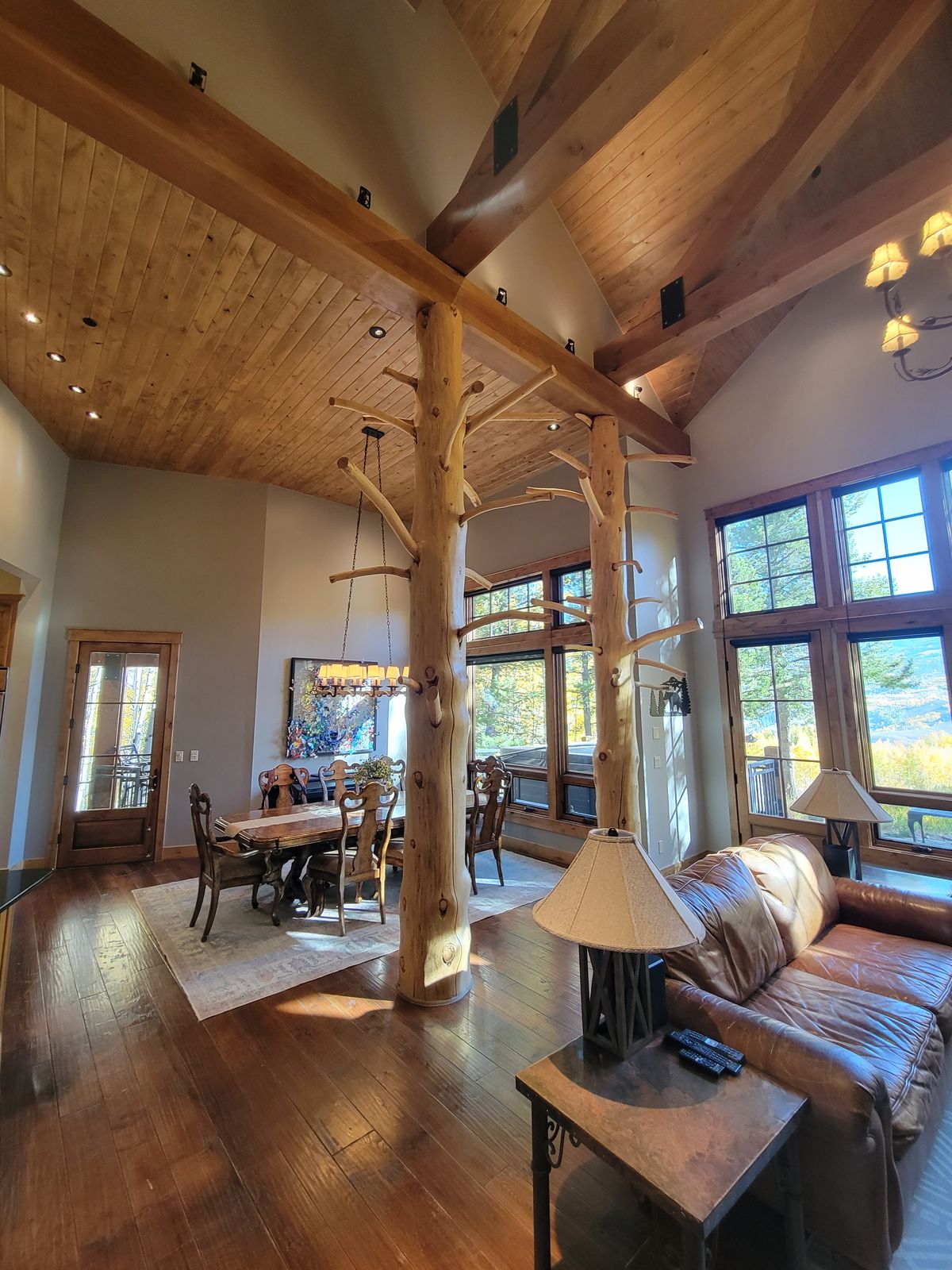 Open great room with vaulted wood-beam and tongue-and-groove ceiling, natural log post columns, hardwood floors, leather sofa, and a dining area visible in the background beside large multi-pane windows.