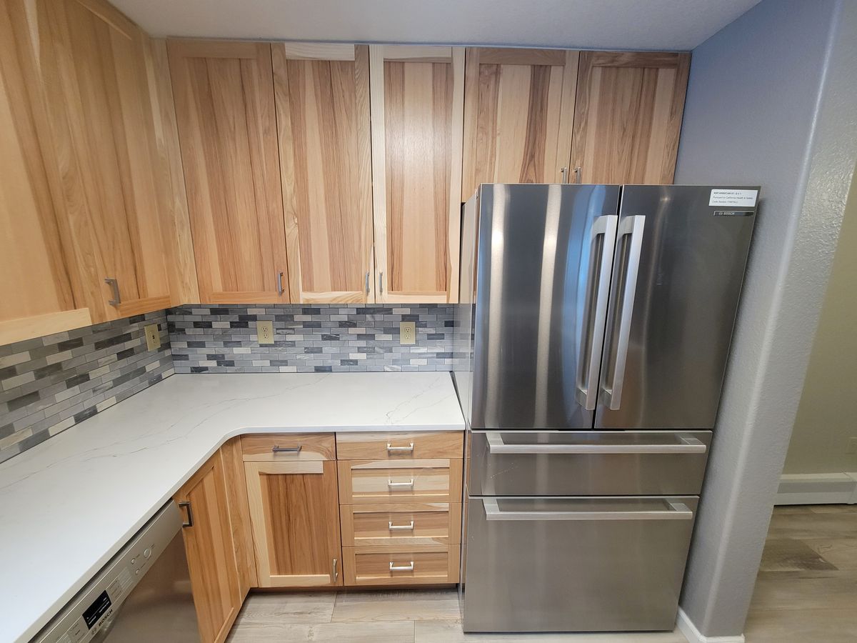 Finished kitchen corner showing light wood upper and lower cabinets, white countertop, mosaic tile backsplash, and a stainless steel French-door refrigerator.