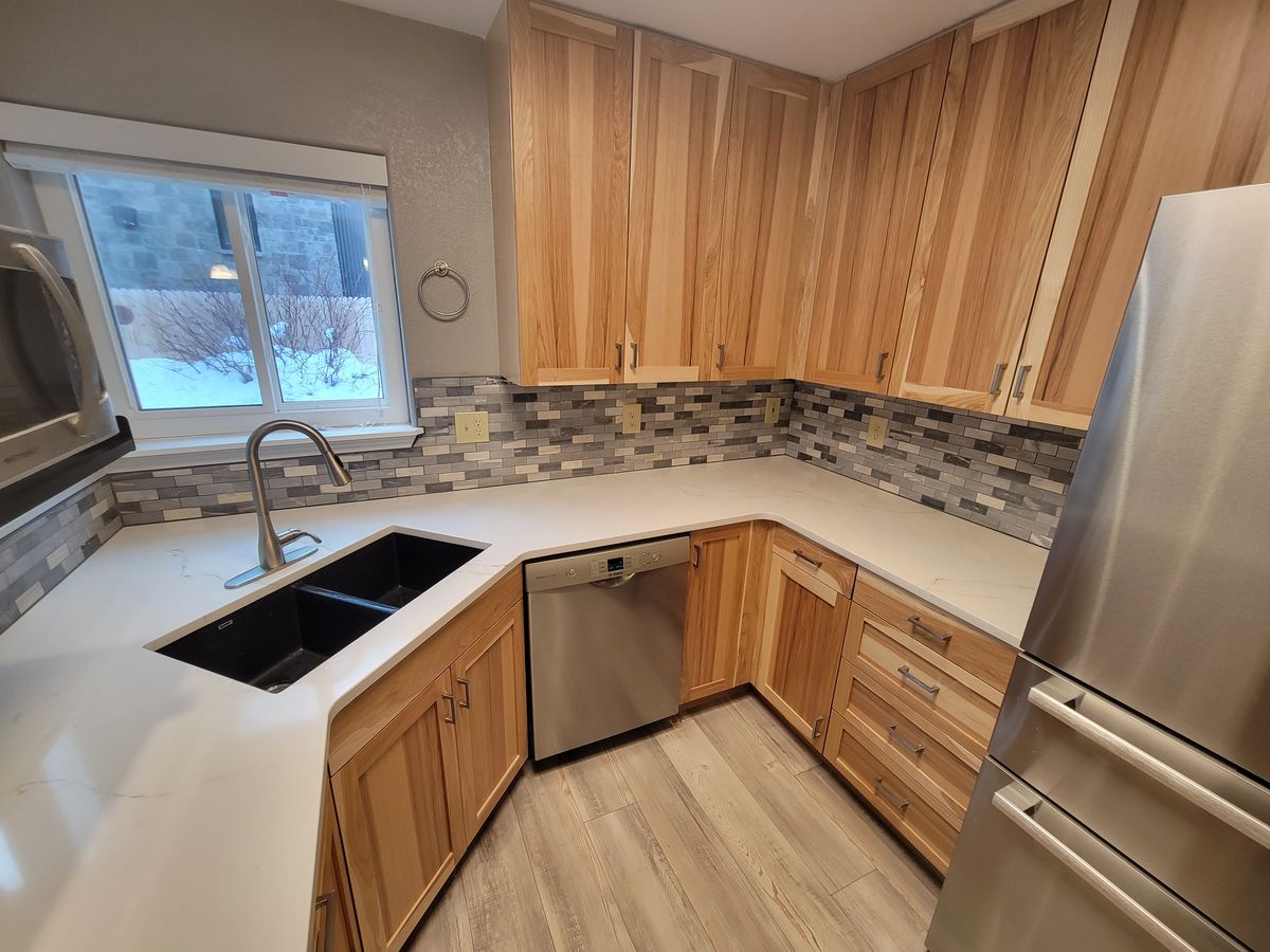 Finished kitchen with light wood cabinets, white countertop, mosaic tile backsplash, black undermount sink, stainless dishwasher, and a window above the sink.