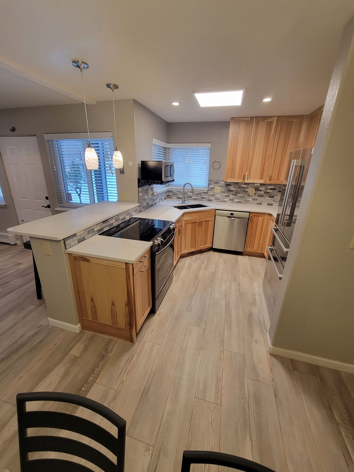 Wide-angle view of finished kitchen with peninsula island, pendant lights, light wood cabinets, mosaic tile backsplash, stainless appliances, and wood-look plank flooring.