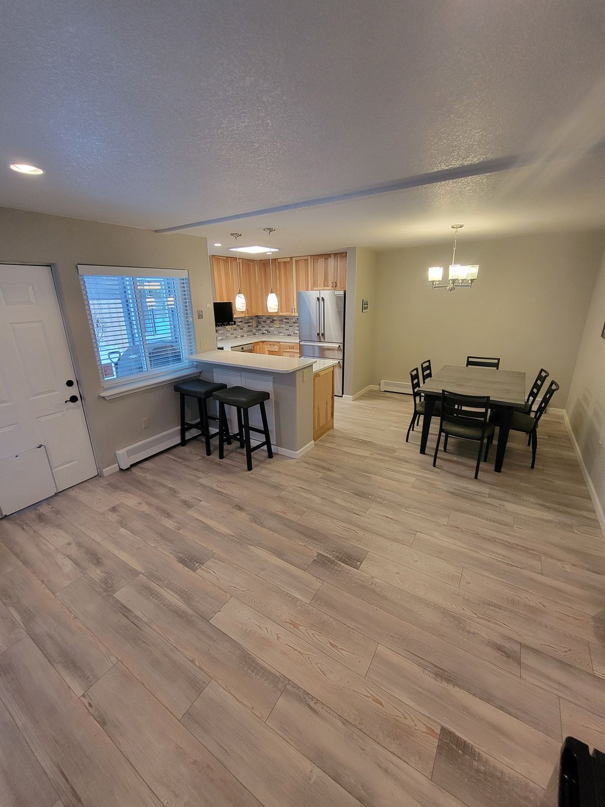 Open-plan view showing the kitchen peninsula with bar stools in the foreground and a dining table with chairs and a chandelier in the background, all on continuous wood-look plank flooring.
