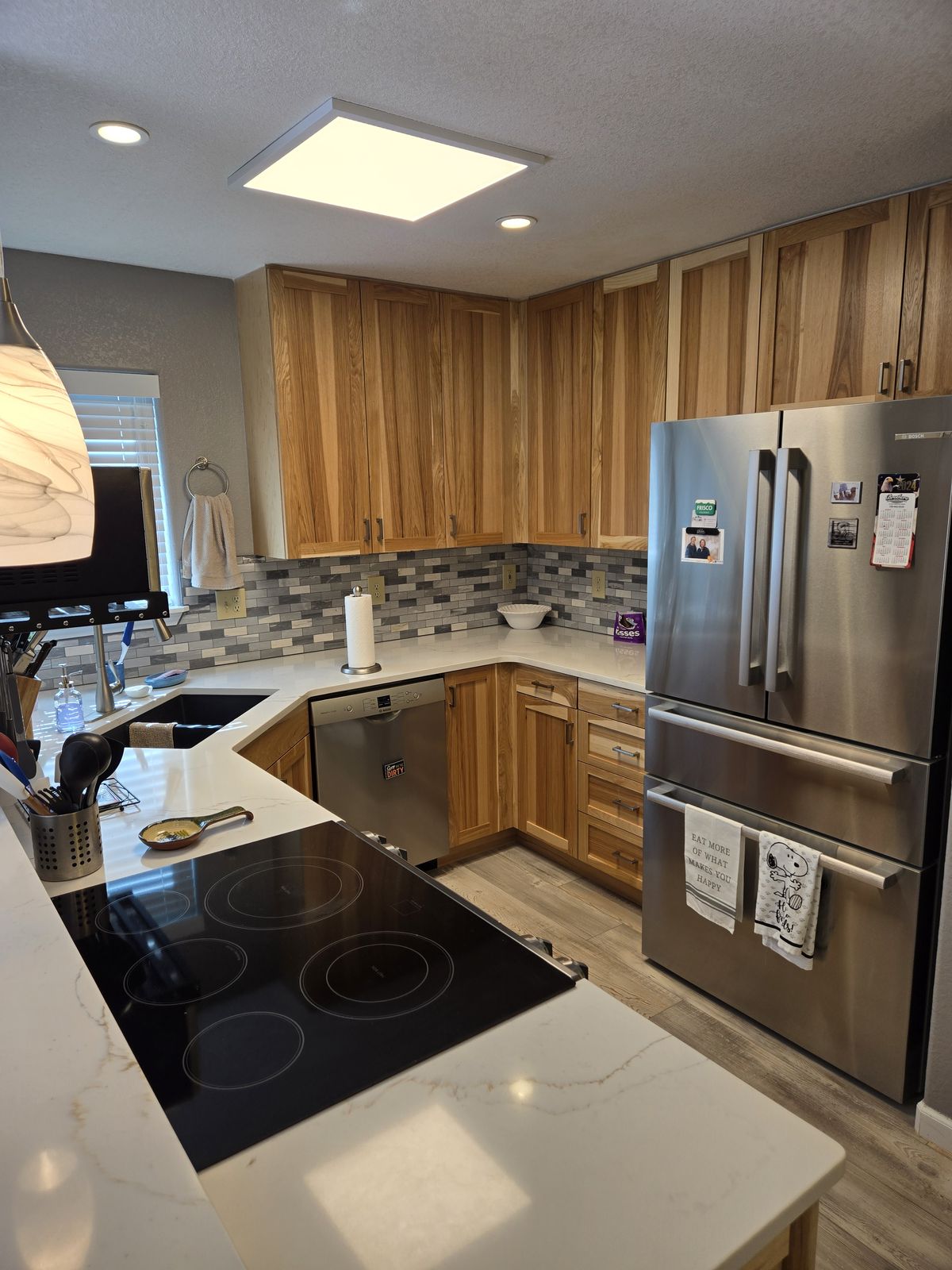 Finished kitchen with light hickory cabinets, white stone countertops, a tile backsplash, stainless appliances including a French-door refrigerator and dishwasher, an electric cooktop on the island, and a skylight above.
