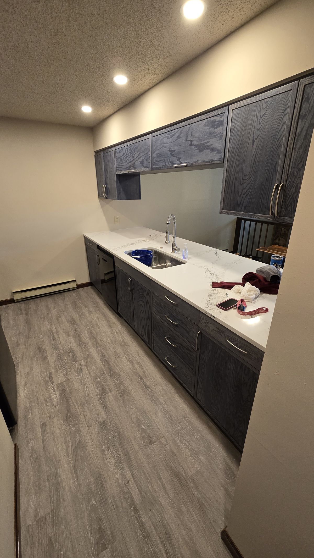 Finished kitchen run showing dark gray upper and lower cabinets, a white veined countertop with undermount sink and faucet, and wood-look plank flooring installed, with a few loose items left on the counter.