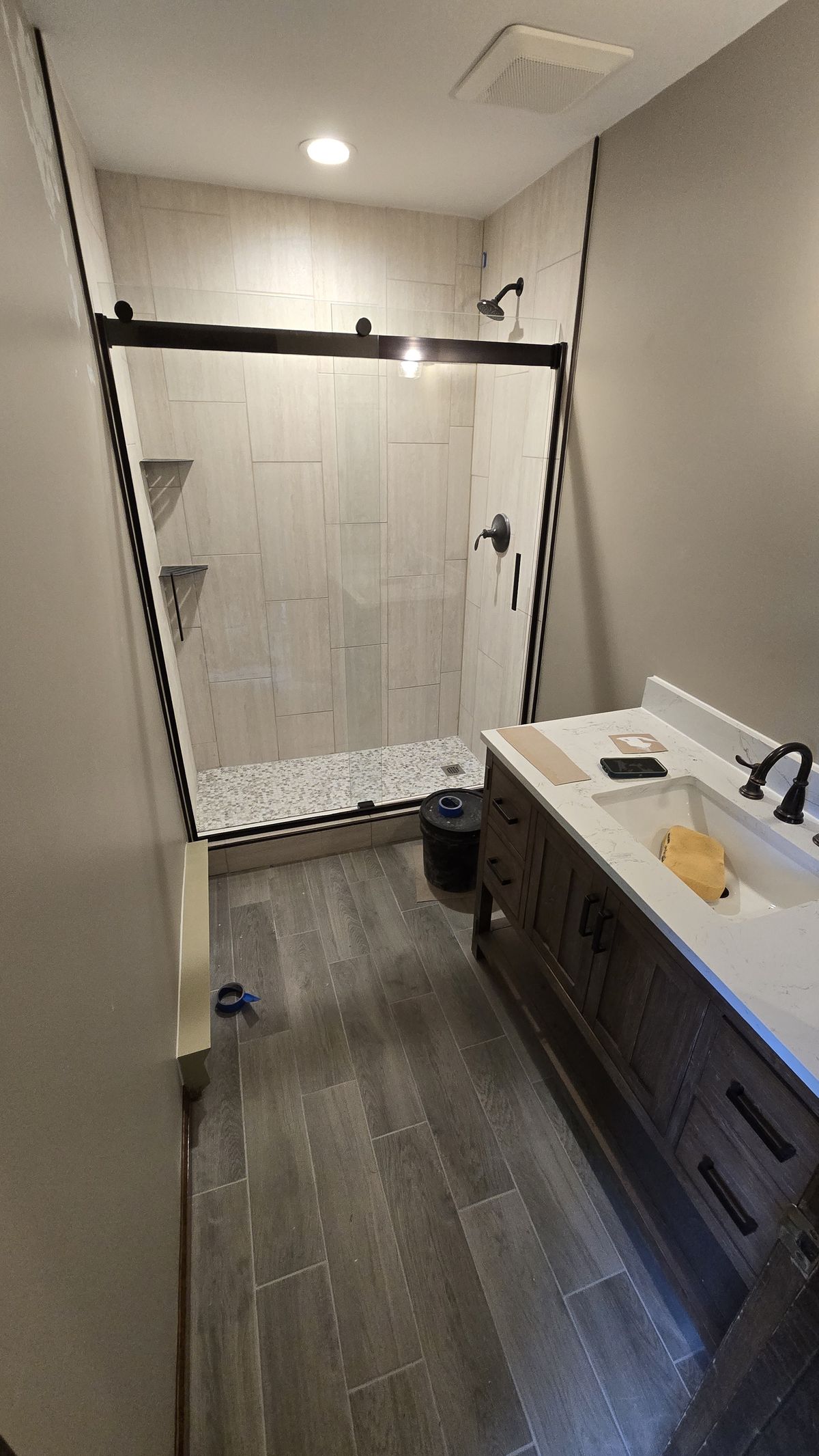 Finished bathroom with a walk-in tile shower enclosed by a black-framed sliding glass door, large white wall tiles and pebble shower floor, wood-look tile flooring throughout, and a dark-cabinet vanity with white countertop.
