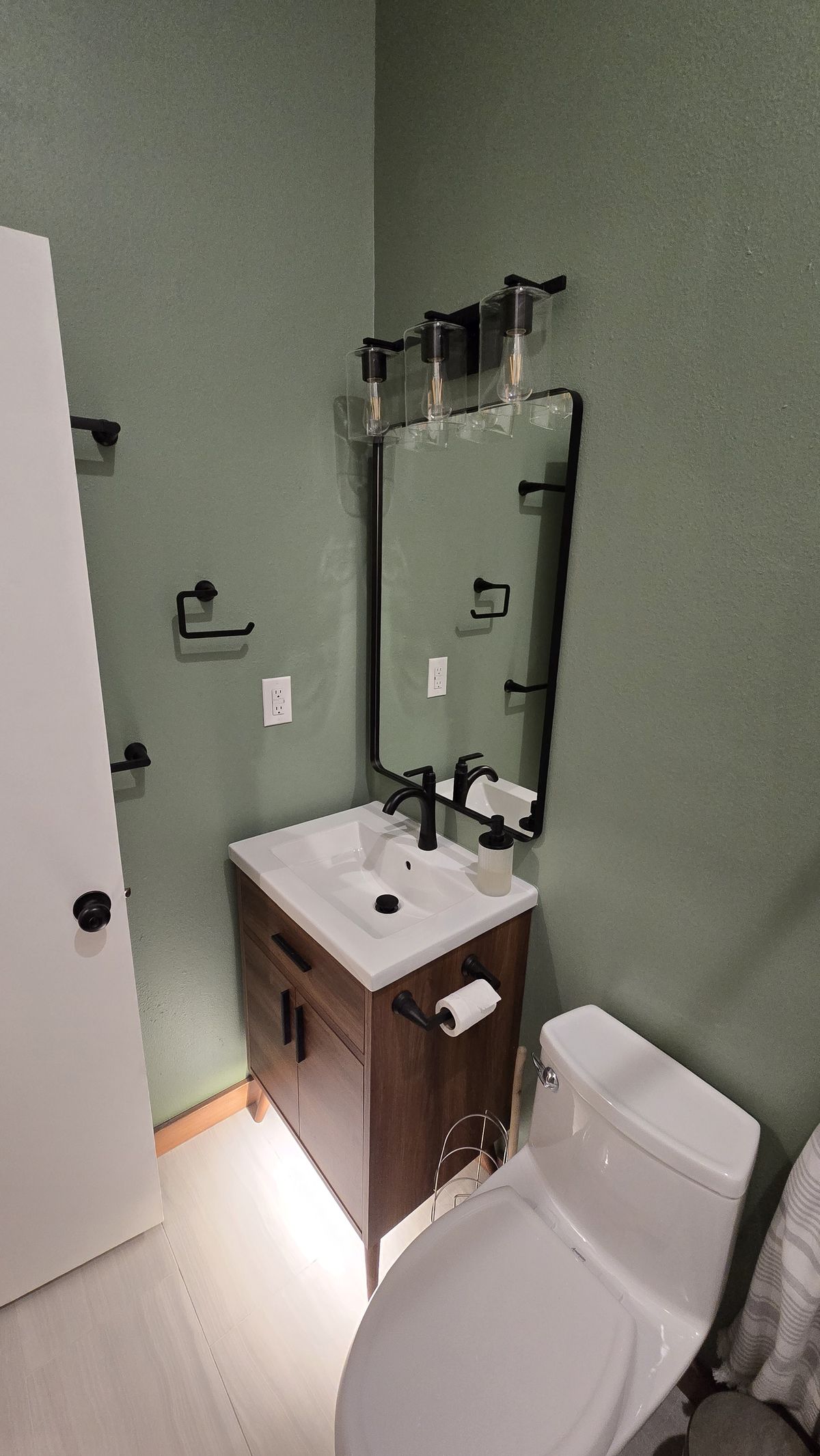 Finished bathroom vanity area with sage-green walls, a wood cabinet vanity with white basin sink, black matte fixtures, a vertical rectangular mirror, a three-bulb black light bar above, and a toilet partially visible to the right.