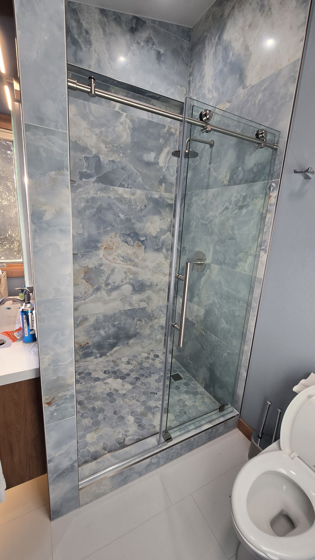Finished walk-in shower with a frameless sliding glass door, large-format blue-gray marble-look tile surround, hexagonal mosaic floor, and a toilet and vanity edge visible alongside.