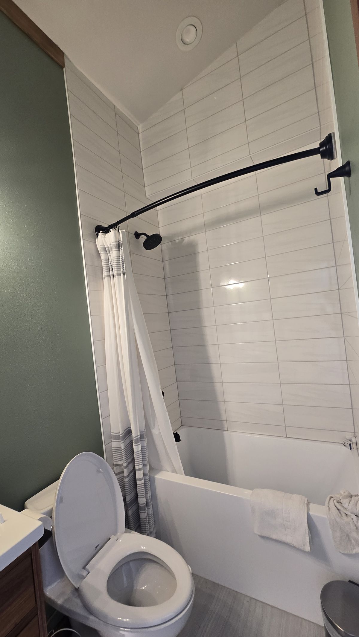 Finished tub-shower combo with white rectangular tile surround and a curved black shower rod with a white curtain, toilet in the foreground with the lid open, and the edge of a wood vanity visible on the left against a sage-green wall.