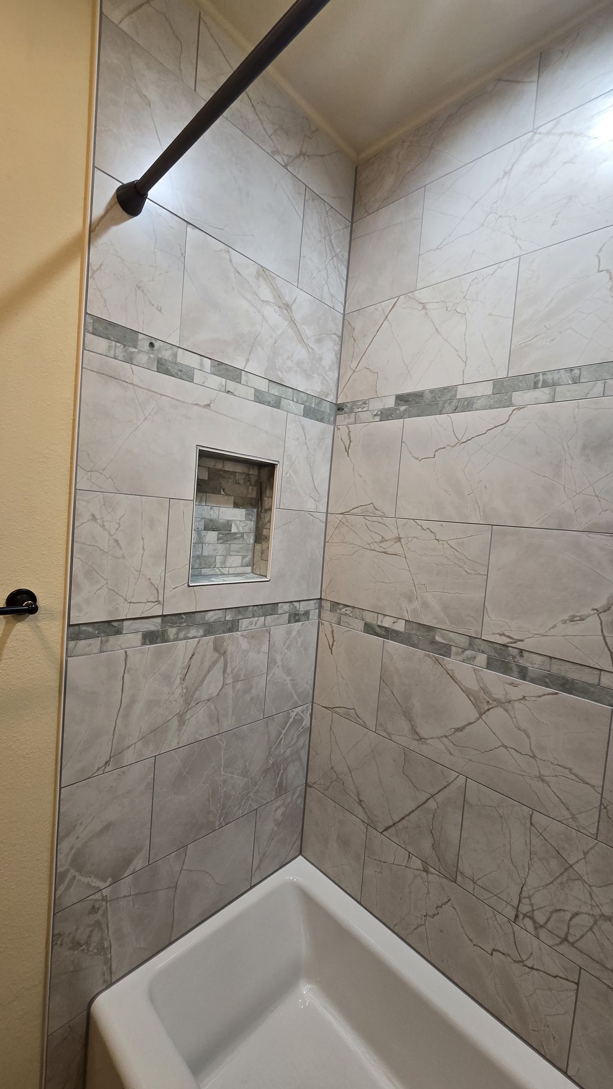 Finished tub/shower surround with large marble-look tiles, a horizontal mosaic border strip, and a recessed mosaic-tiled niche, bathtub visible at the bottom.