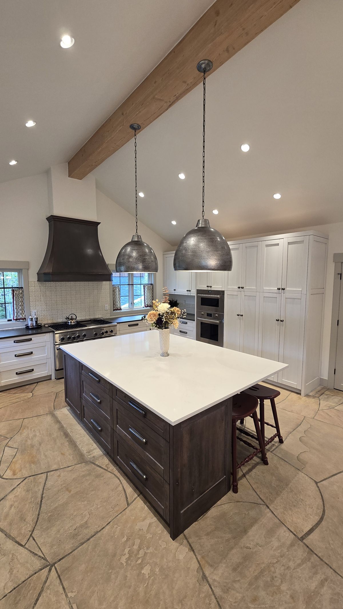 Finished kitchen viewed from the island end showing the dark island with white countertop, custom range hood, white upper and lower cabinets, pendant lights, and flagstone tile floor.