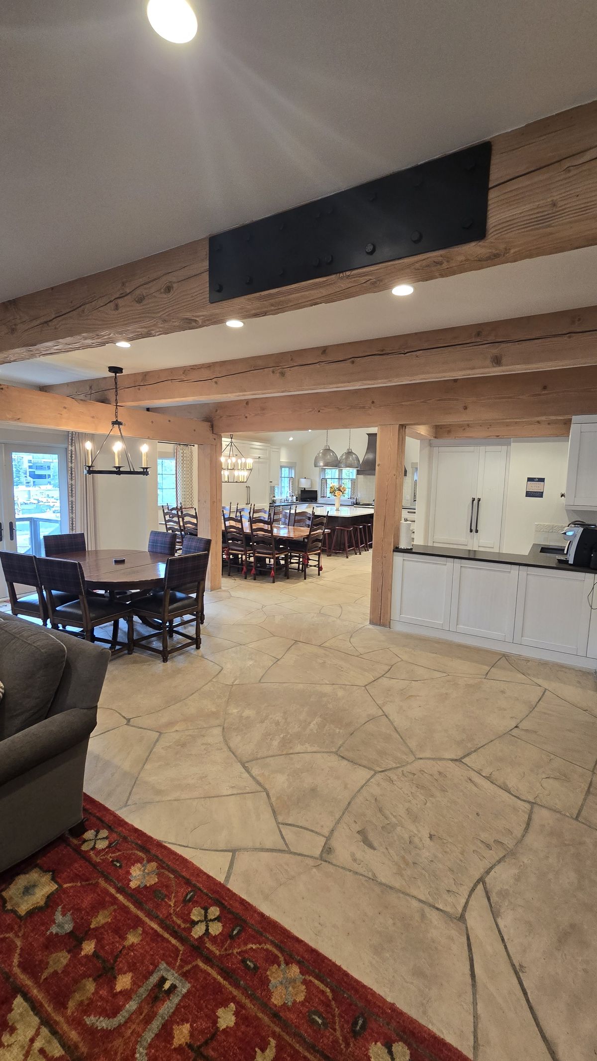 Finished open-plan living, dining, and kitchen area with exposed wood post-and-beam structure, large-format flagstone tile floor, dining table with chairs, and kitchen visible in the background.