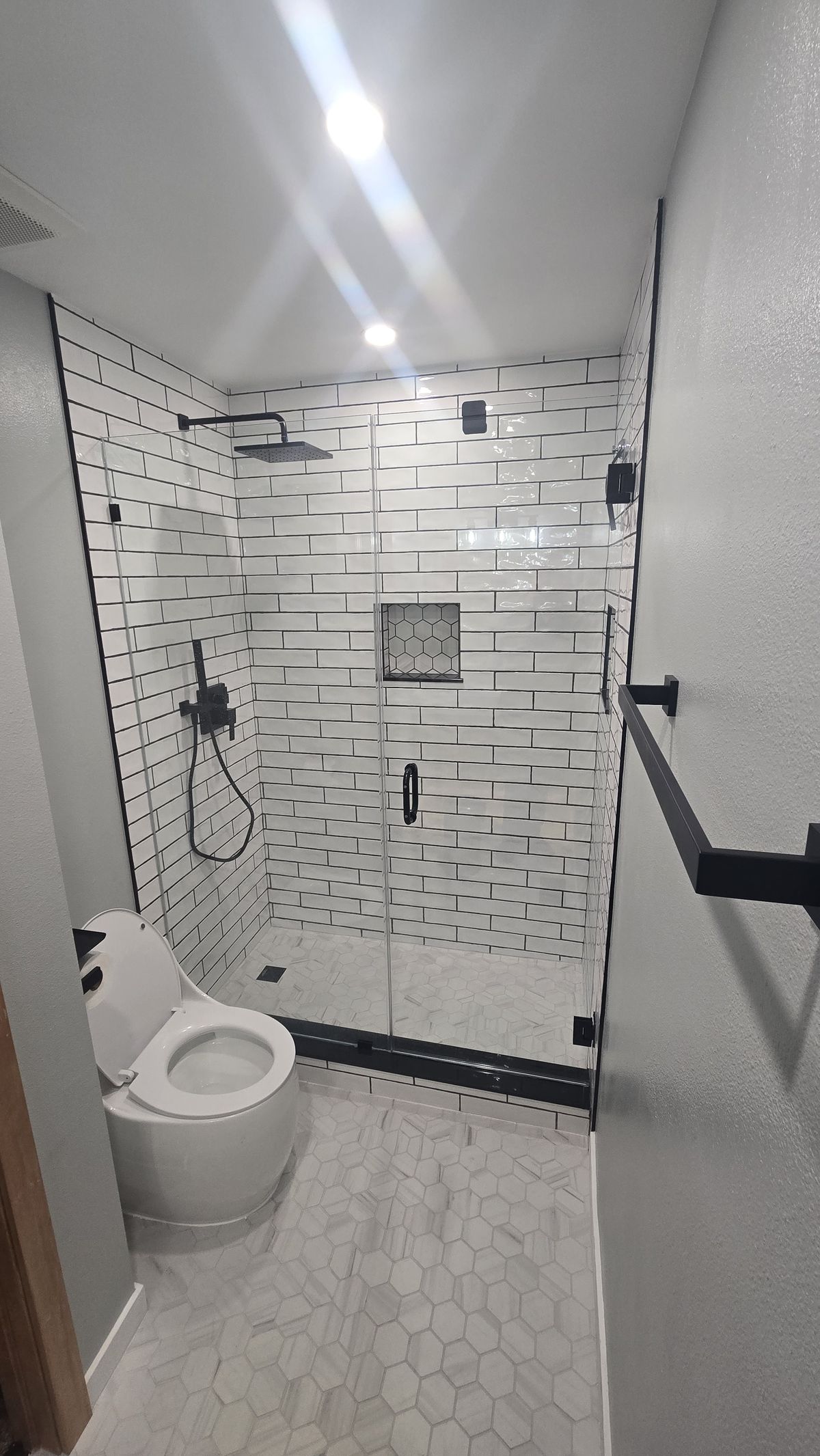 Finished bathroom with a frameless glass shower enclosure, white subway tile with dark grout on shower walls, a built-in niche, black fixtures, hexagon floor tile, and a toilet visible.