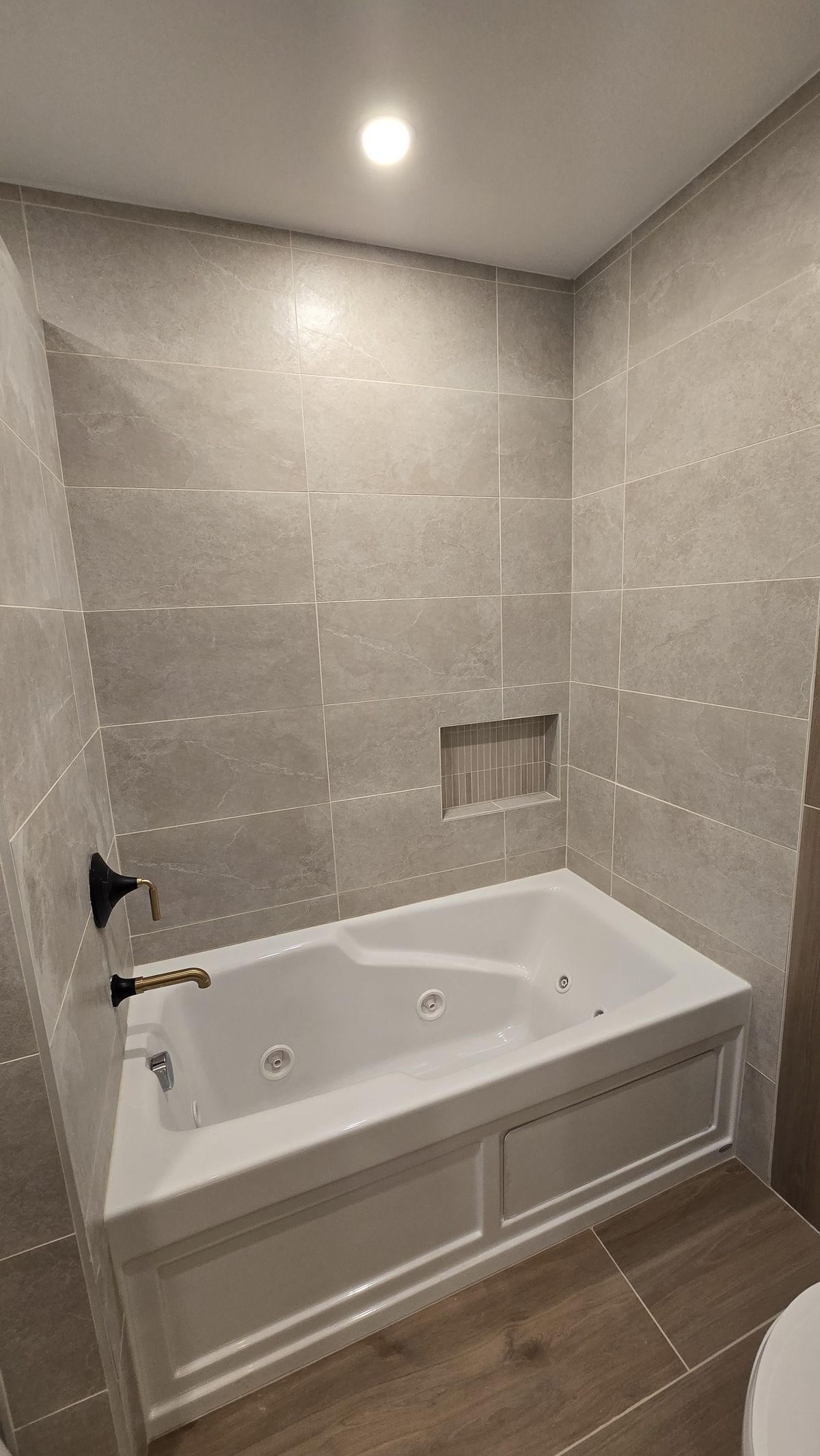 Finished jetted soaking tub set into a large-format tile surround with a recessed niche and matte-black tub filler, recessed lighting above.