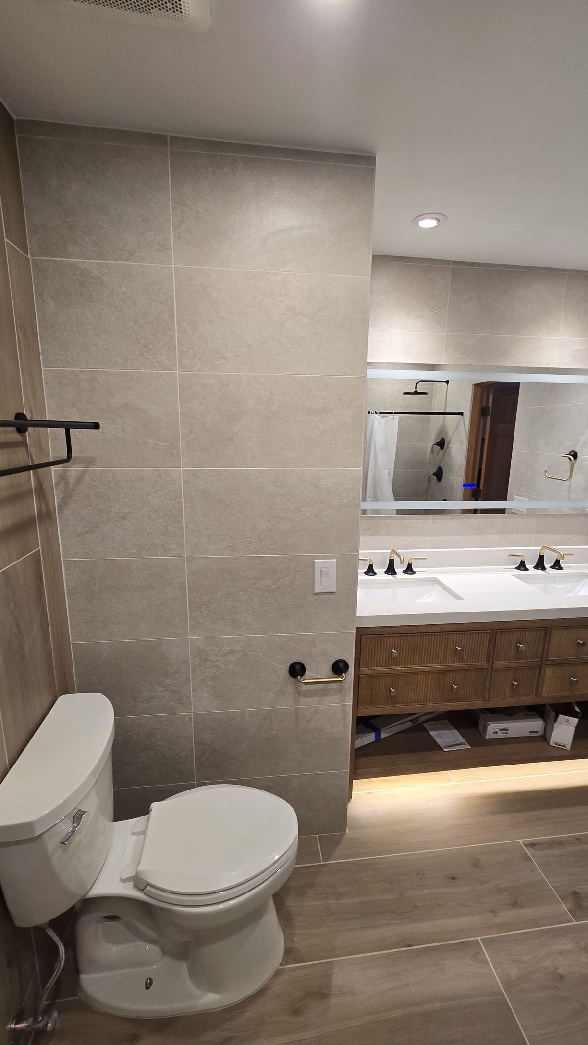 Finished bathroom with large-format tile walls, wood-look tile floor, toilet, and double-sink vanity with wood-tone base and large frameless mirror; no people or debris visible.