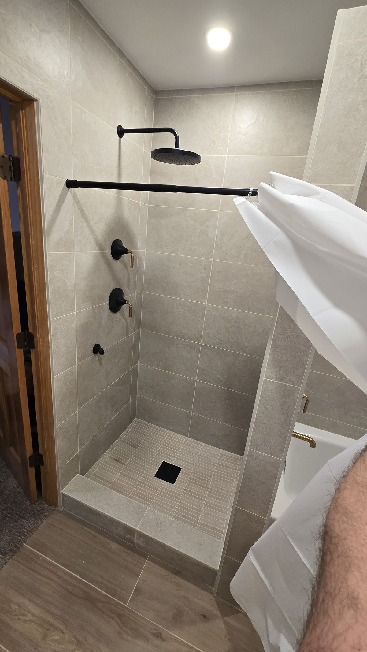 Finished walk-in shower with large-format gray wall tile, matte black rain head and control valve, black square drain, and wood-look tile floor, with a white curtain partially in frame at right.