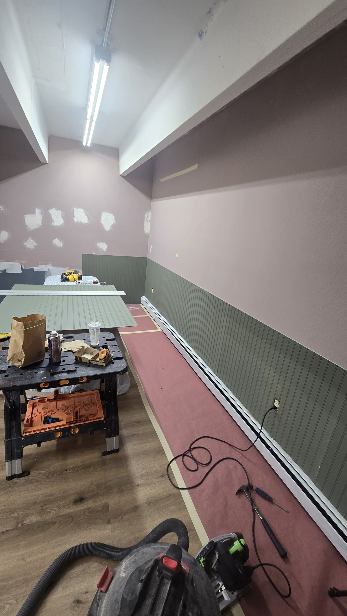 Long room under construction with pink-painted upper walls, green beadboard wainscoting, LVP flooring partially protected by red paper, fluorescent strip lighting, and a portable workbench with tools.